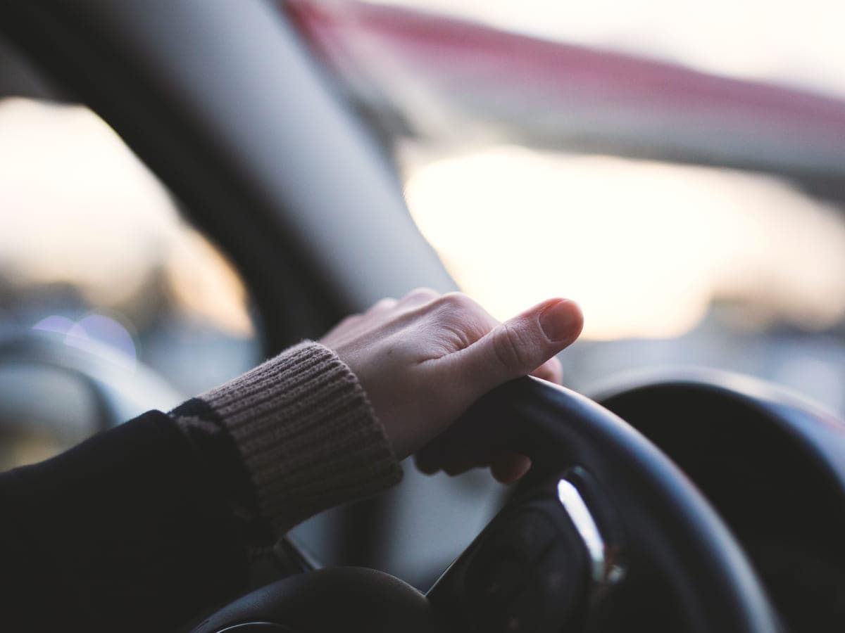 A hand resting lightly on a car steering wheel, knitted jumper cuff, soft overcast daylight through the windscreen